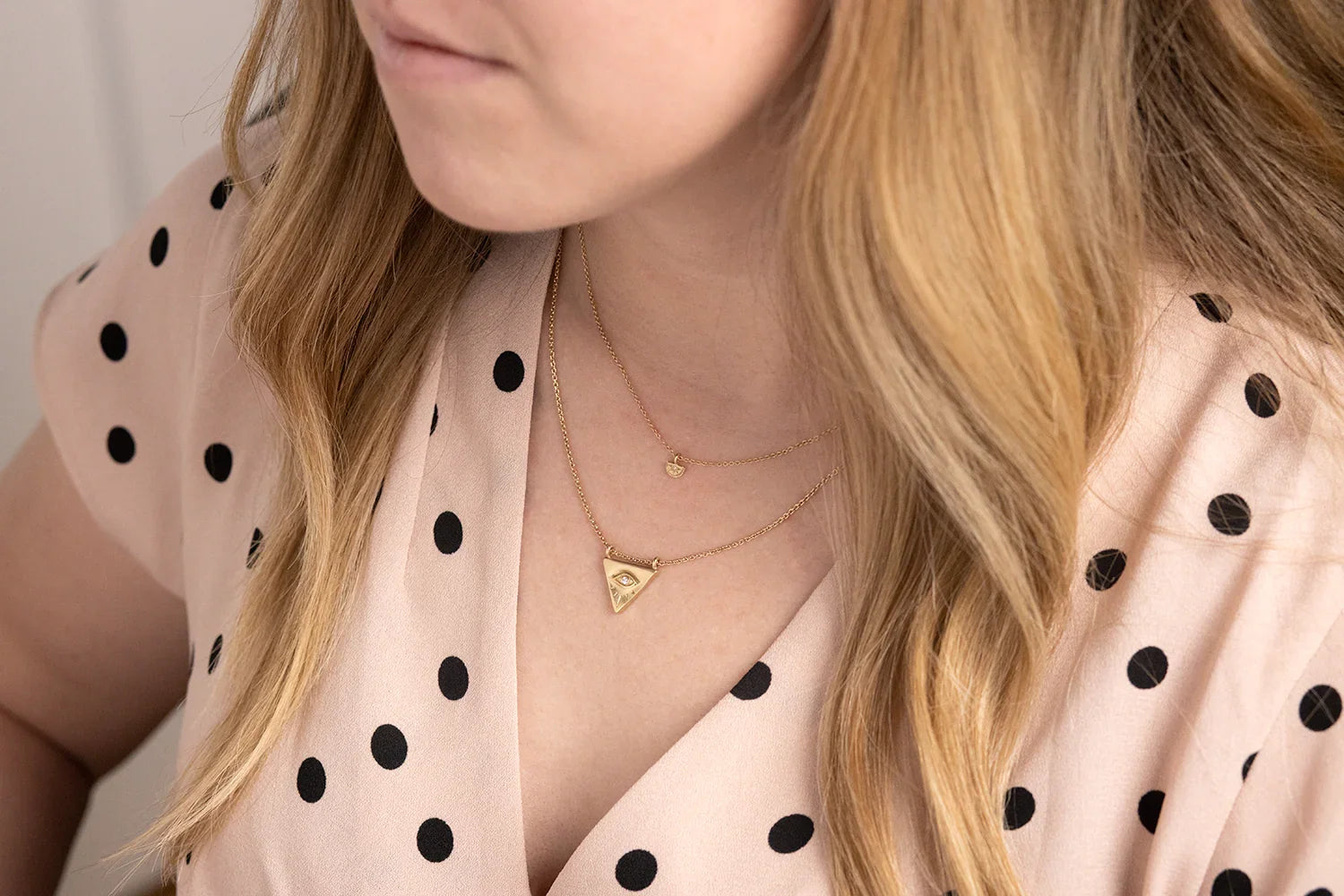 Close-up of woman wearing layered 18k gold Evil Eye pendant necklaces, soft natural light, beige polka dot blouse