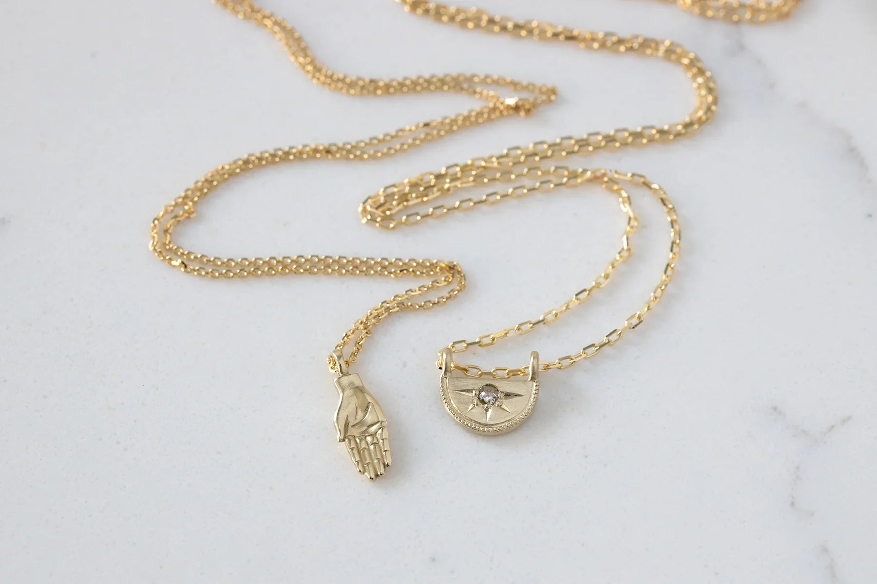 Elegant Fairmined gold hand and celestial pendants on delicate chain, natural light on white stone
