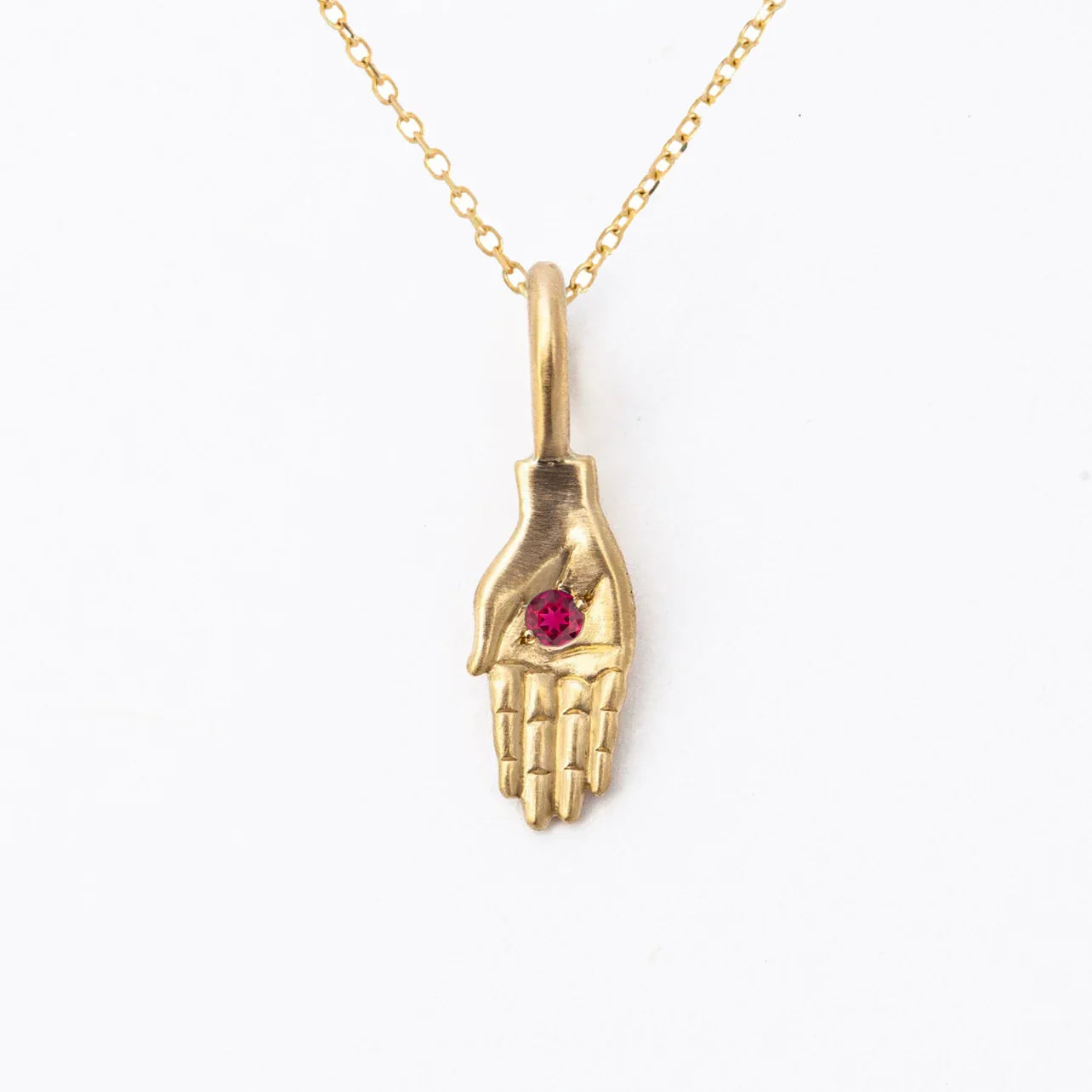 Handcrafted gold hand pendant with deep red gemstone on delicate chain, minimalist white background