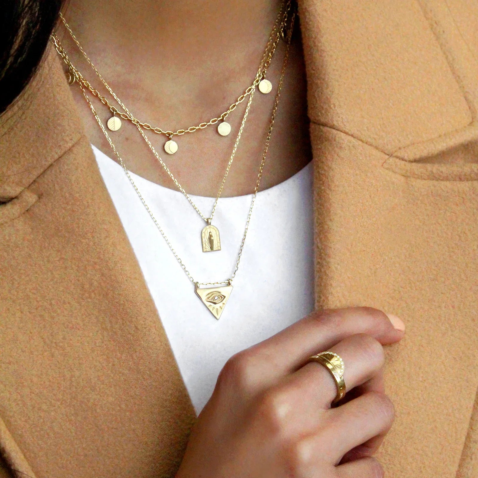 Layered Fairmined gold necklaces and ring with sun and eye motifs, warm natural light on neutral clothing