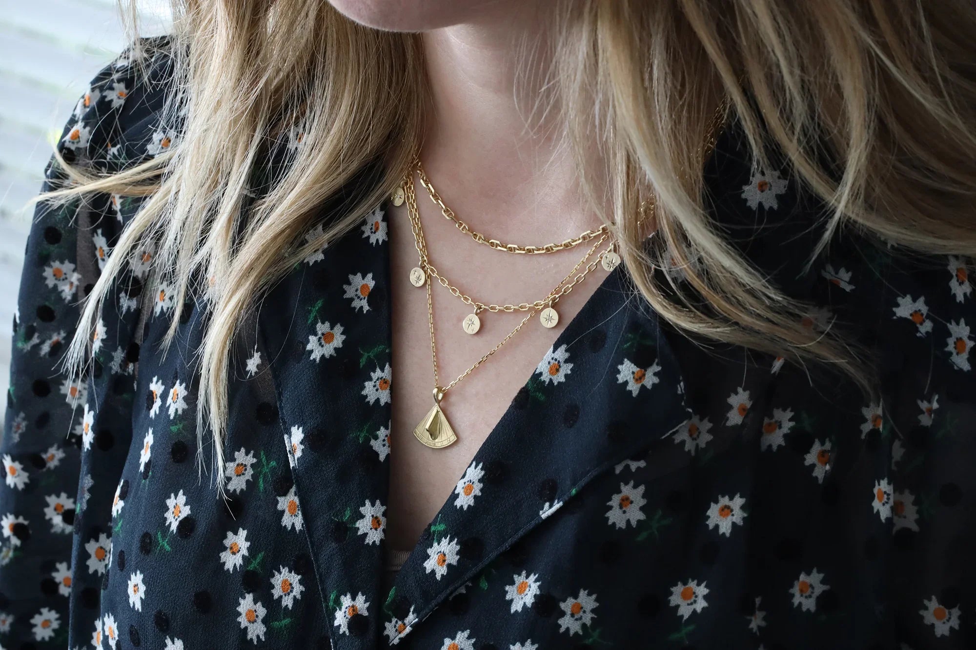 Close-up of layered Fairmined gold sundial pendant necklace and charm chain on woman wearing floral black blouse in soft natural light
