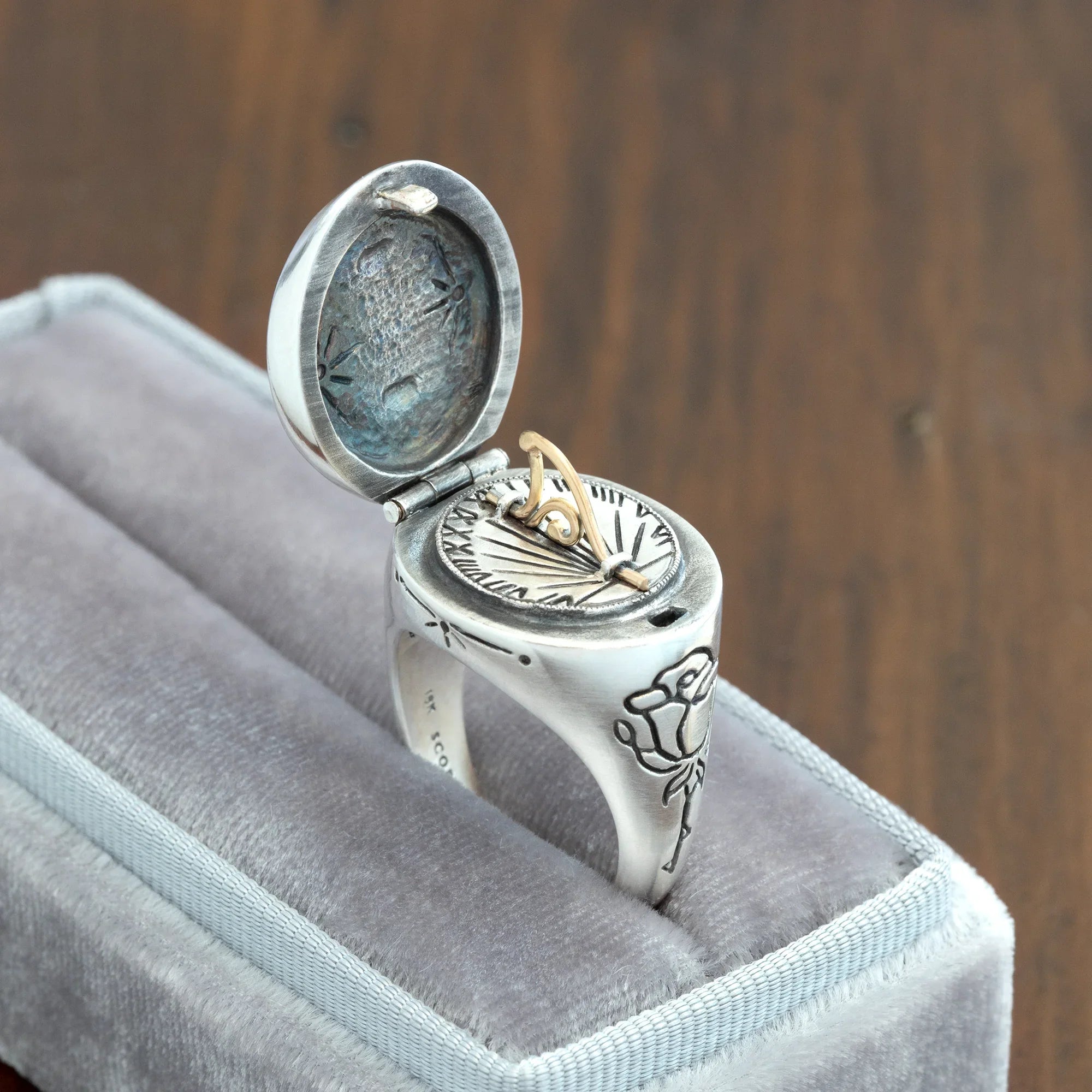Handcrafted silver sundial ring with intricate rose engraving, gold gnomon dial, displayed in grey velvet box on wood