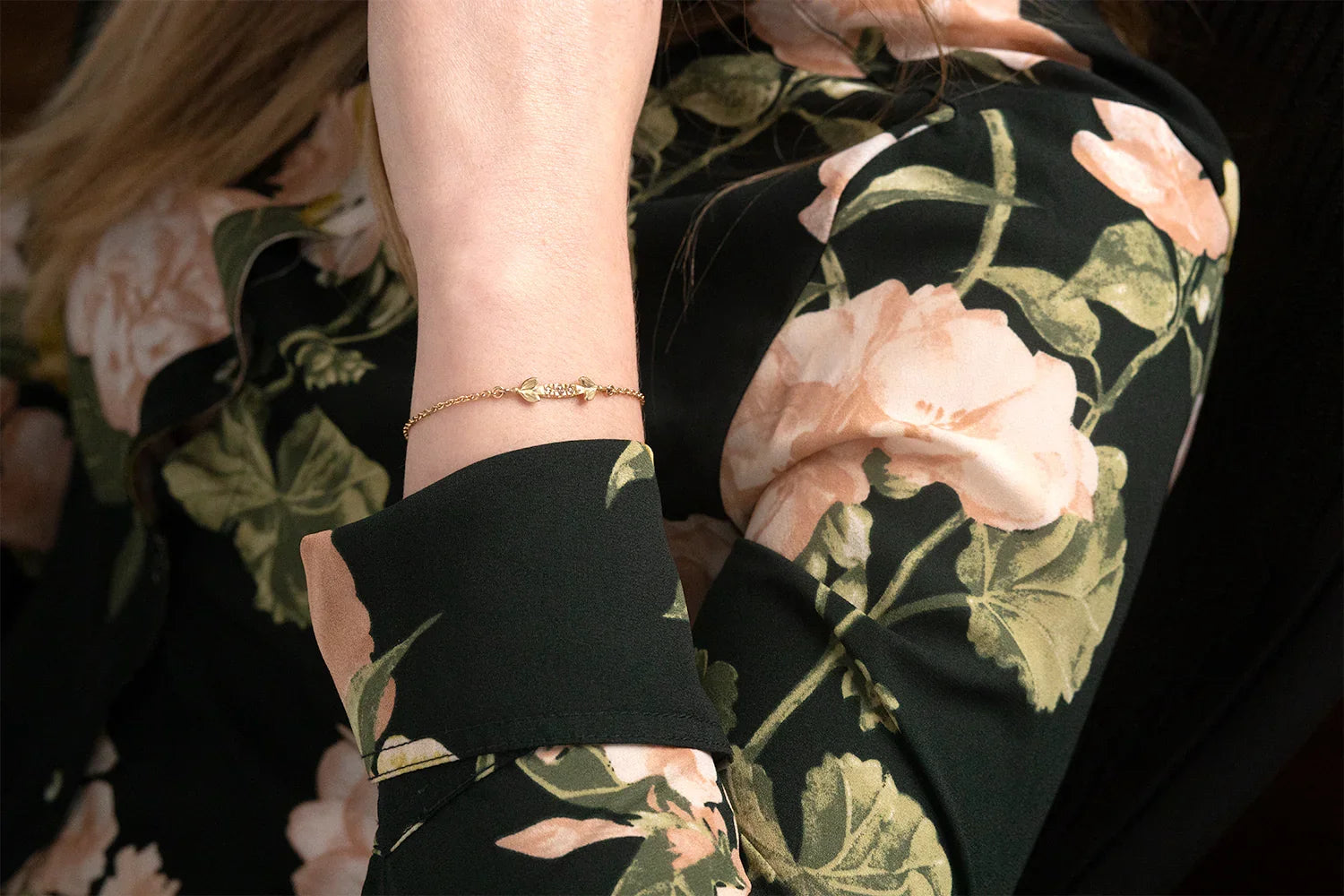 Delicate handcrafted 14k gold bracelet with leaf motifs on wrist, natural light, floral blouse background