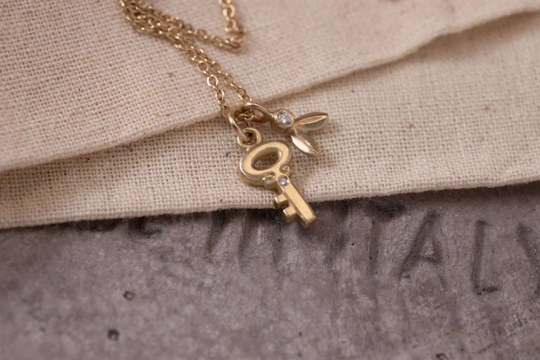 Close-up of delicate 14k gold key and leaf charms with small diamonds on chain, natural linen and stone background