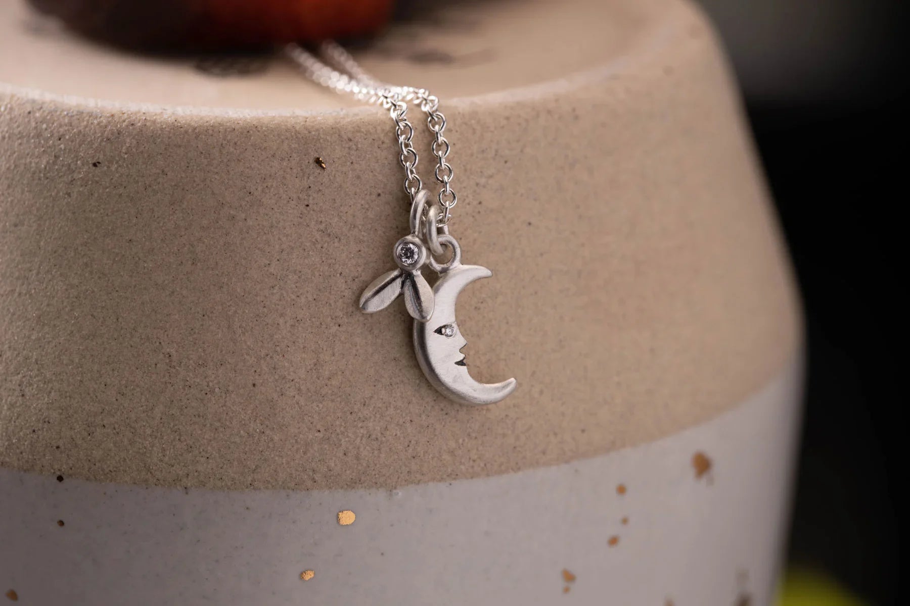 Close-up of silver crescent moon pendant with diamond eye and flower charm on chain, minimalist ceramic background