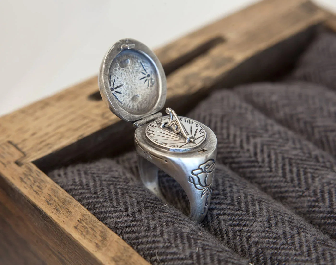 Close-up of silver sundial ring with hinged lid and engraved floral details on textured fabric in wooden box