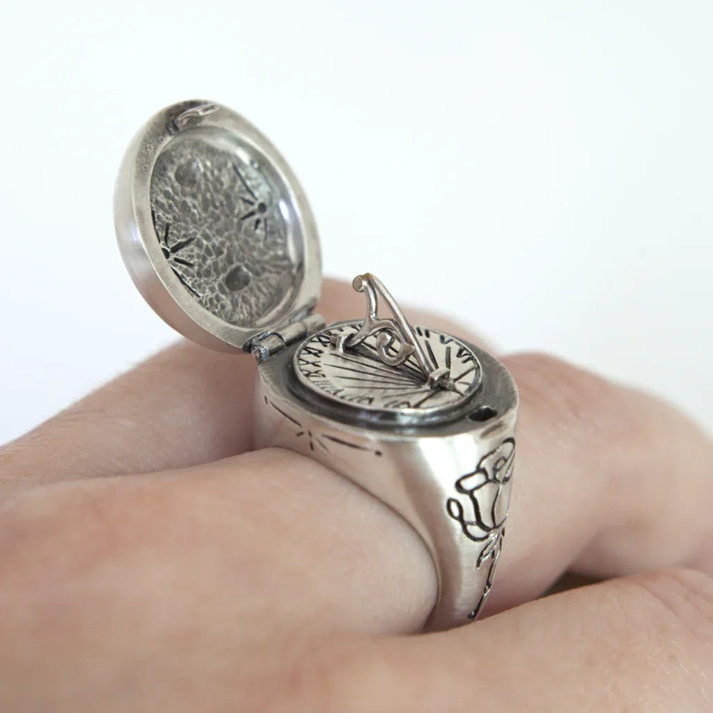 Close-up of silver sundial ring with open lid showing engraved hour markers and floral design on finger