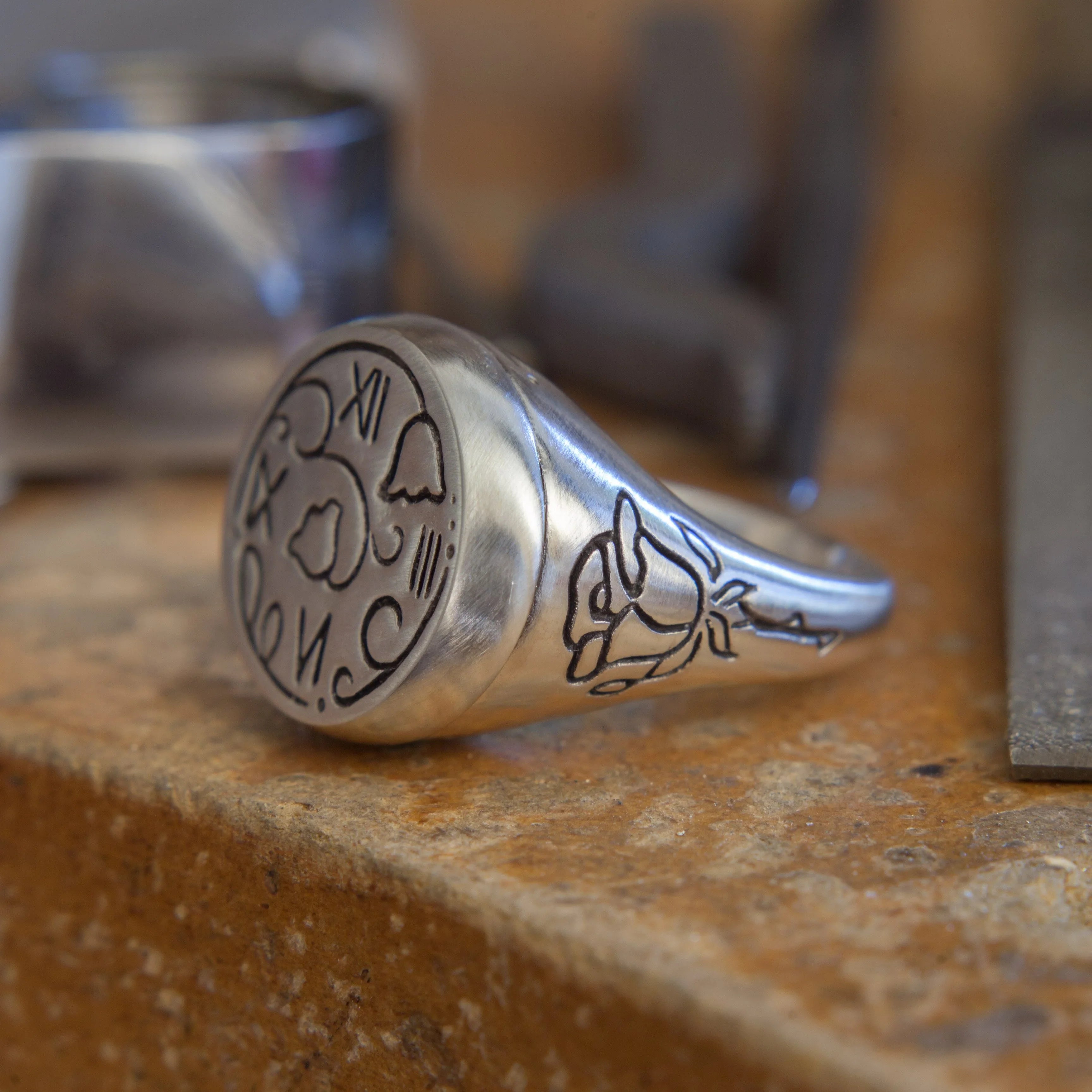 Close-up of silver signet ring with hand-engraved floral and clock face motifs on stone surface