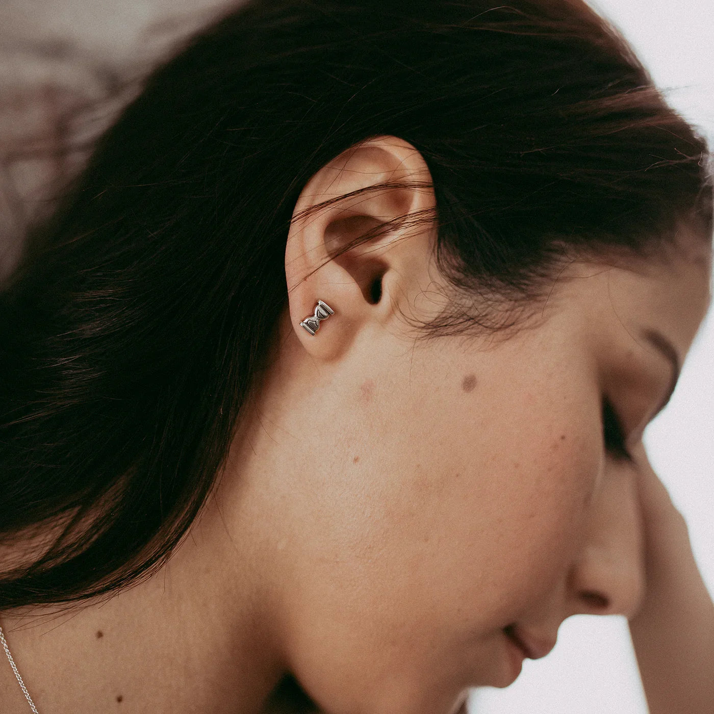 Close-up of woman wearing elegant silver hourglass stud earring, natural soft lighting, minimalist background