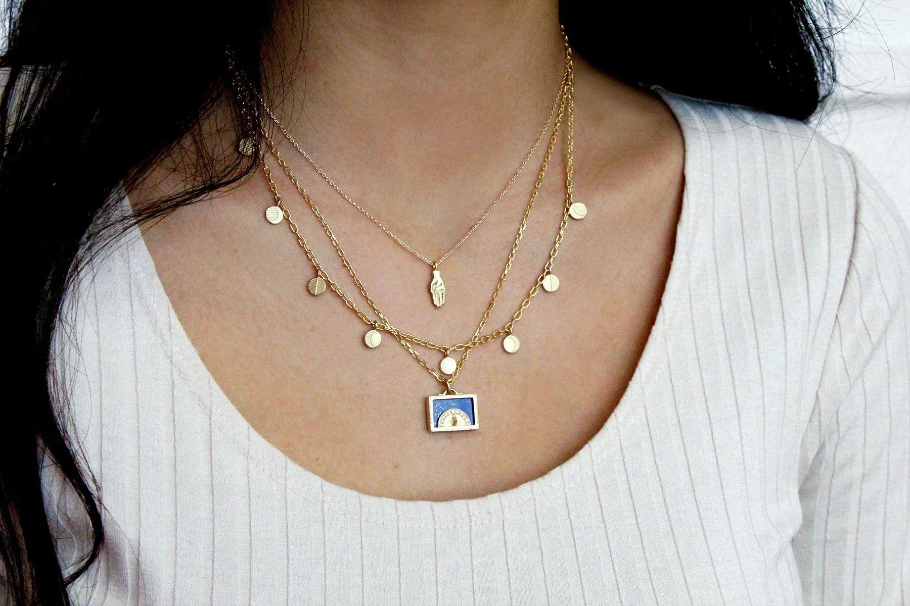 Layered gold necklaces with sundial pendant, discs, and hand charm on woman's neck, soft natural light