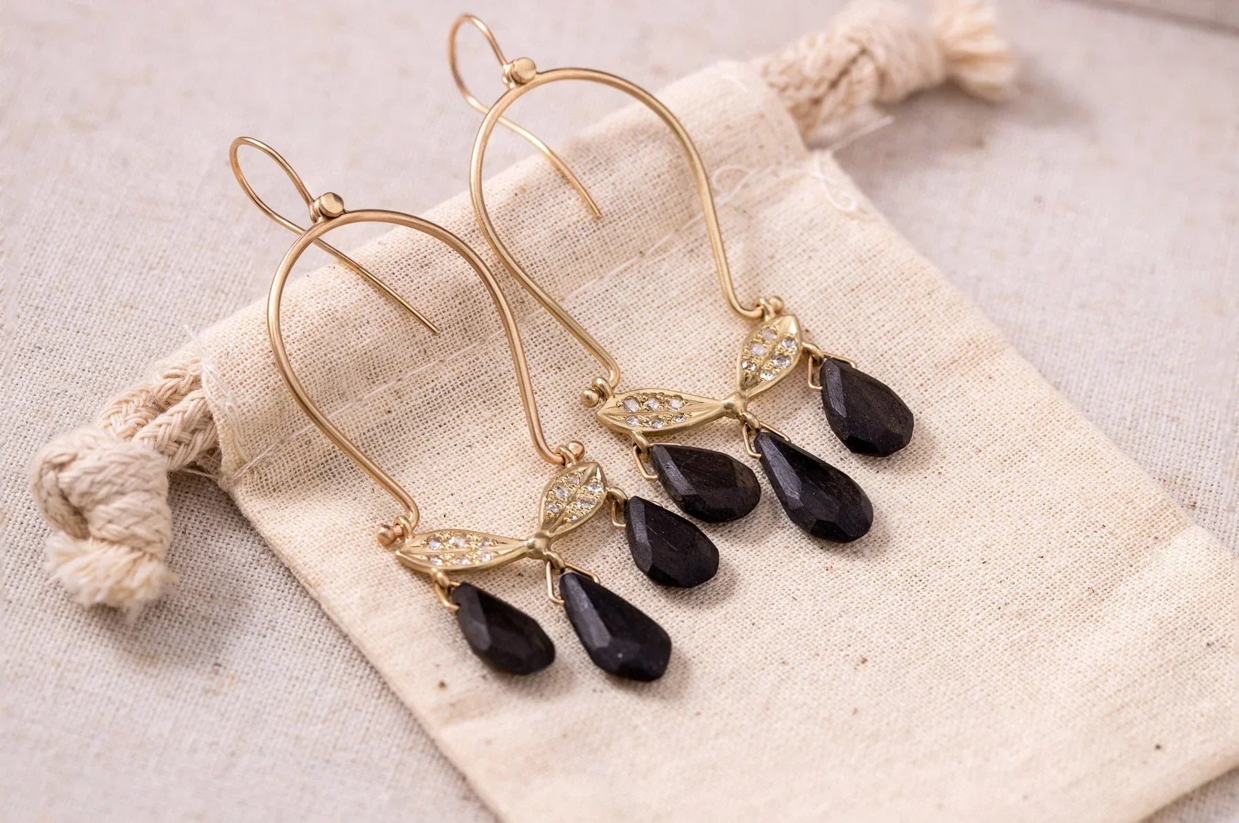 Elegant handcrafted gold earrings with small crystals and faceted black teardrop stones on natural linen bag, warm soft light