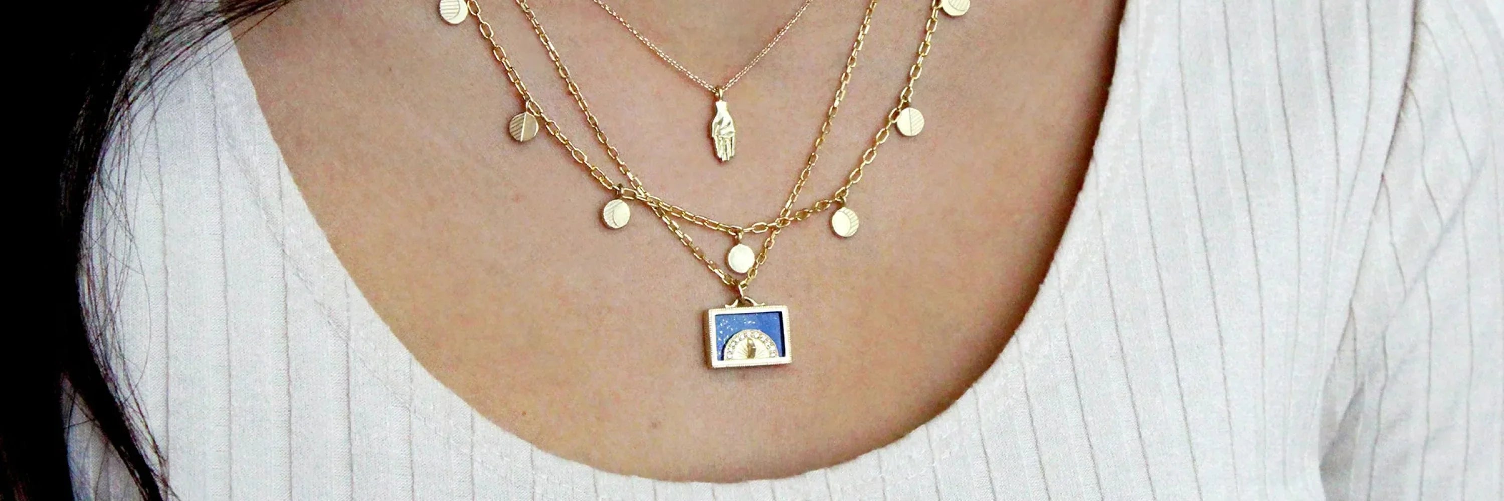 Close-up of layered Fairmined gold necklaces with sundial pendant and hand charm on neutral ribbed top