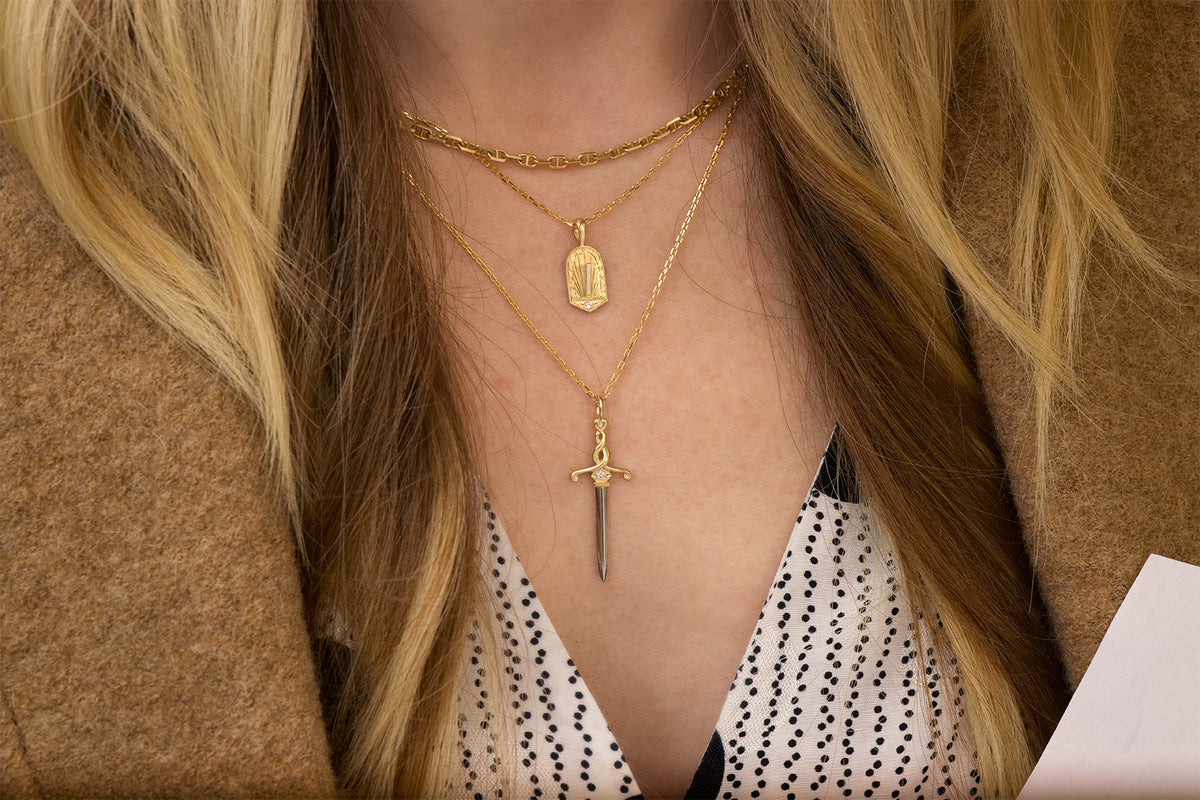 A close-up of a person wearing layered gold necklaces, including a yellow gold sword pendant with a platinum blade and white diamond set into the handle. The other pendant has the design of a small vertical sundial with white diamond set into the base and milgrain border. The person in the photo is also wearing a choker length gold mariner chain. They are styled with a brown coat and a black-and-white polka dot top.